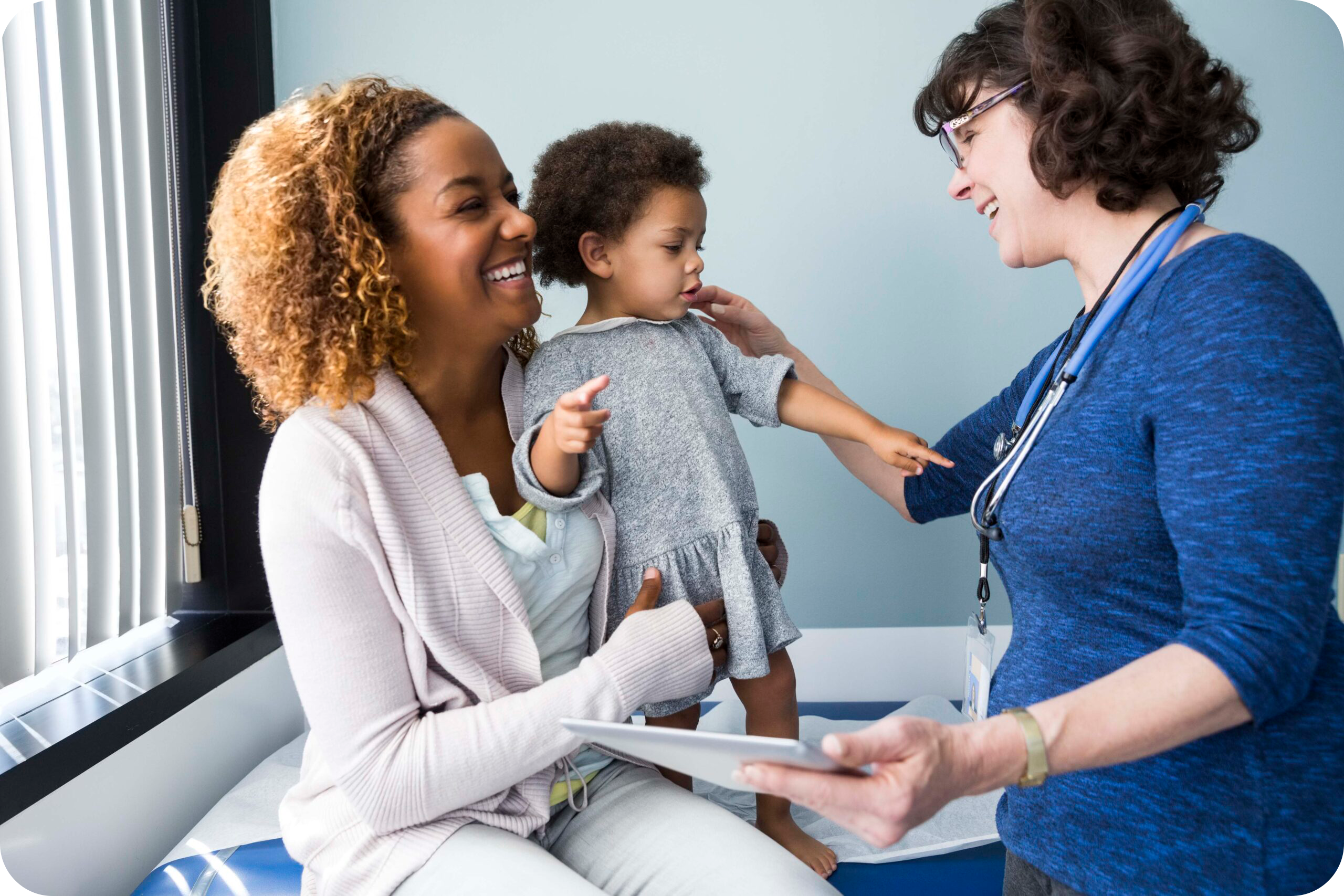 Smiling White female pediatrician greets a toddler and her Black mother in an exam room.